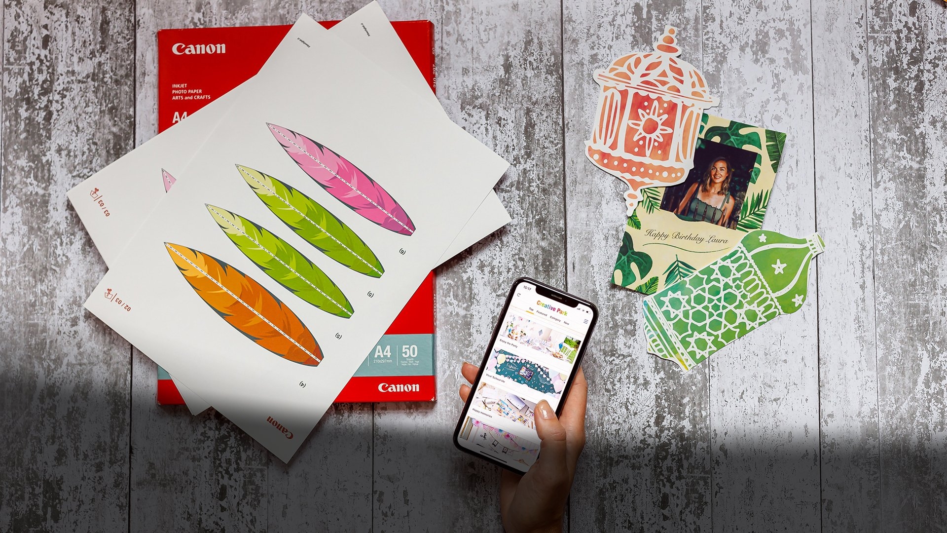 A hand holding a smartphone displaying the Creative Park app. Canon printer paper and Creative Park templates and designs are arranged on the table.